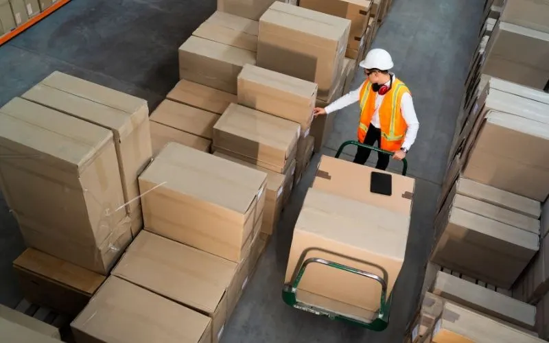 A warehouse worker in a hard hat and safety vest moving boxes on a cart, seen from above